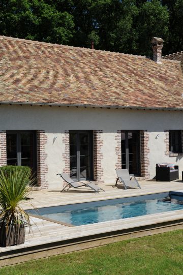 Les portes vitrées et leur encadrement de briques traditionnel rythme la longère en arrière plan de la piscine aux tons naturels.